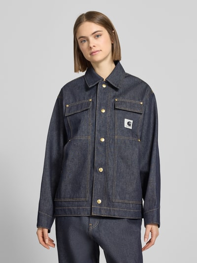 Frauenmodel trägt Regular Fit Jeansjacke mit Logo-Patch Modell 'Arctic' von Carhartt Work In Progress aus dunkelblauer Baumwolle, sichtbare Nähte, Umlegekragen, Knopfleiste, Brust- und Eingrifftaschen, Nahaufnahme Oberkörper.