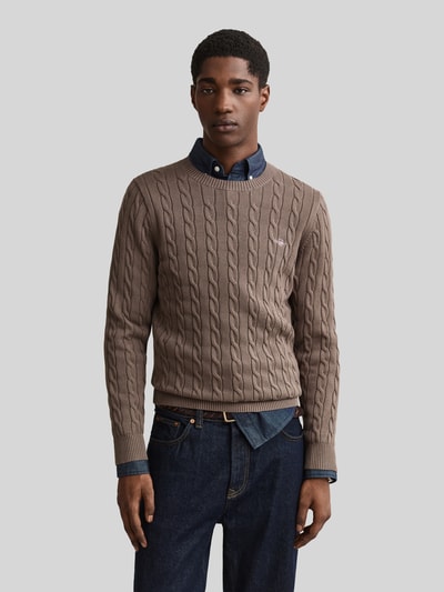 Männermodel trägt braunen Regular Fit Pullover aus reiner Baumwolle von GANT, Zopfmuster, kombiniert mit Jeans, Ganzkörperansicht von vorne.