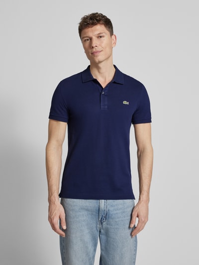Męski model nosi Slim Fit Poloshirt aus reiner Baumwolle von Lacoste, efekt: jednolity kolor, krój slim fit, wyhaftowane logo, kołnierzyk polo, rękaw: krótki rękaw, zakończenie rękawów: prążkowane, wykończenie: z rozcięciami po bokach, wykończenie materiału: pika, bawełna, Ganzkörperansicht von vorne.