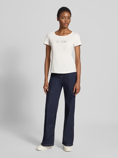 Kobiece modelka nosi Damski t-shirt Regular Fit T-Shirt aus reiner Baumwolle von Tom Tailor w kolorze kremowym, gładka tekstura, proste cięcie, granatowe spodnie, zdjęcie całej sylwetki od przodu.