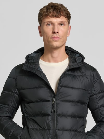 Męski model nosi Steppjacke mit Kapuze und Reißverschluss von MCNEAL, czarny kolor, pikowana faktura, krój regular fit, zbliżenie na górną część ciała, długość tyłu 70 cm, szerokość ramion w rozmiarze M 46 cm, szerokość w klatce piersiowej w rozmiarze M 54 cm, długość rękawa w rozmiarze M 63 cm.