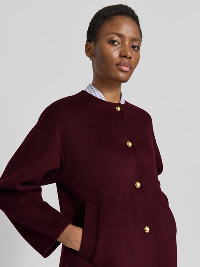 Frauenmodel trägt Relaxed Fit Kurzmantel aus Woll-Mix mit Kaschmir-Anteil von GANT, unifarben in dunklem Bordeauxrot, Struktur glatt, seitliche Eingrifftaschen, einreihige goldfarbene Knöpfe, Rundhalsausschnitt, langarm, Nahaufnahme Oberkörper.