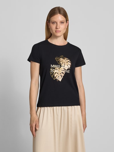 Kobiece model prezentuje damski t-shirt z bawełny od Soyaconcept, model 'RINA', krój regular fit, okrągły dekolt, nadruk z napisem i motywem, długość tyłu w rozmiarze S: 60 cm, czarny, złote i beżowe detale, gładka tekstura, widok na całe ciało z przodu.