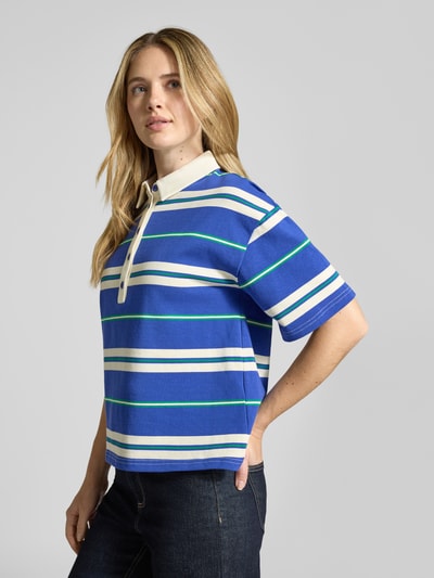 Frauenmodel trägt Loose Fit Poloshirt mit überschnittenen Ärmeln Modell 'Ulma' von Lollys Laundry aus reiner Baumwolle, breite blaue und weiße Streifen mit grünen Akzenten, Polokragen, Rückenlänge bei Größe S 58 cm, Ganzkörperansicht von vorne.