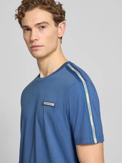 Männermodel trägt T-Shirt mit Label-Patch und Rundhalsausschnitt von EA7 Emporio Armani in Blau, körpernah geschnitten, Nahaufnahme Oberkörper.
