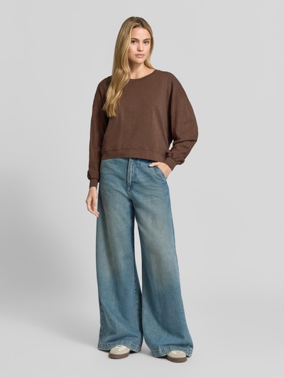 Frauenmodel trägt Relaxed Fit Sweatshirt aus reiner Baumwolle von American Vintage, braun, elastische Bündchen, Rundhalsausschnitt, langärmlig, kombiniert mit weiter Jeans, Ganzkörperansicht von vorne, Rückenlänge bei Größe S 57 cm.