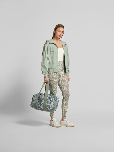 Frauenmodel trägt Regular Fit Jacke mit Logo-Stitching von Lala Berlin in hellem Grün, Kapuze mit Tunnelzug, Eingrifftaschen, Reißverschluss, kombiniert mit gemusterten Leggings, Ganzkörperansicht von vorne. Rückenlänge bei Größe S: 53 cm, Brustweite 60 cm, Ärmellänge 59 cm.