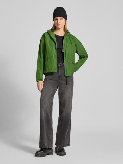 Vrouwenmodel draagt groene Jacke mit Pattentaschen Modell 'MILENA' van Didriksons, recht pasvorm, capuchon, labelprint, ganzkörperaanzicht van voren. Ruglengte maat 36: 60 cm, schouderbreedte: 60 cm, borstwijdte: 55 cm, mouwlengte: 66 cm.