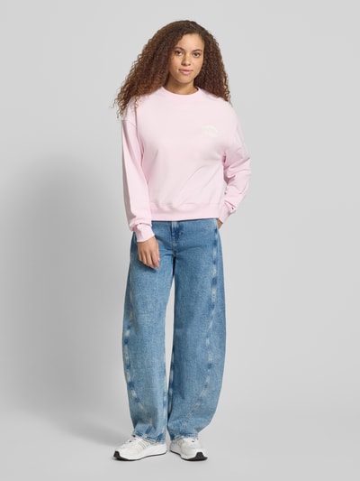 Frauenmodel trägt Boxy Fit Sweatshirt aus reiner Baumwolle von Tommy Jeans, Optik: Unifarben, Label-Print, Crew Neck, Langarm, gerippter Abschluss, Materialverarbeitung: Sweat, Farbe: Hellrosa, Ganzkörperansicht von vorne.
