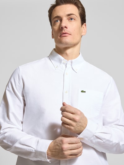 Męski model nosi Regular Fit Freizeithemd aus reiner Baumwolle von Lacoste, efekt: jednolity kolor, detal z logo, koszula casualowa oxford z bawełny, kołnierzyk typu button down, listwa guzikowa, długi rękaw, sportowy mankiet, brystasza, zaokrąglone wykończenie, przód, zbliżenie na górną część ciała.