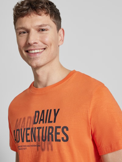 Męski model nosi Regular Fit T-Shirt aus reiner Baumwolle von camel active, pomarańczowy, nadruk z napisem, okrągły dekolt, krój regular fit, długość tyłu w rozmiarze M 68 cm, zbliżenie na górną część ciała.
