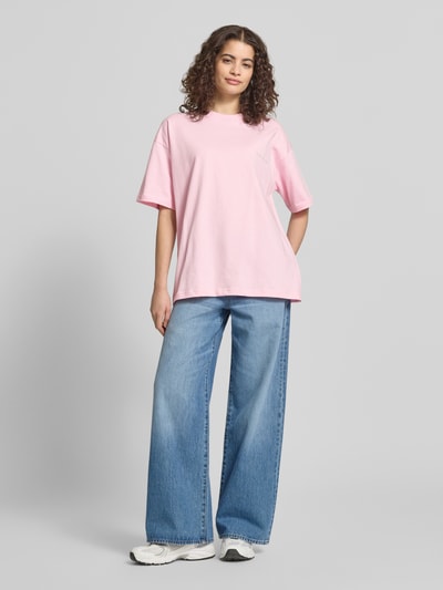 Frauenmodel trägt Damen T-Shirt mit Logo-Stitching von ANOTHER COTTON LAB, Reine Baumwolle, Lockerer-Schnitt, Rundhalsausschnitt, Überschnittene Schultern, hellrosa Farbe, glatte Textur, kombiniert mit blauen Jeans und weißen Sneakers, Ganzkörperansicht von vorne.