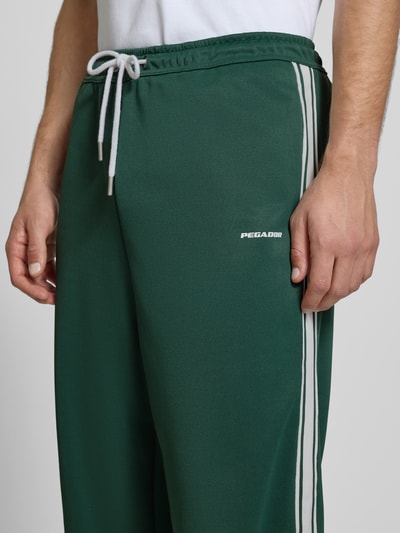 Männermodel trägt Wide Leg Sweatpants mit elastischem Bund von Pegador in Dunkelgrün mit weißen Streifen, Logo und Tunnelzug, Nahaufnahme Oberkörper.