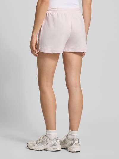 Frauenmodel trägt Straight Fit Shorts mit Tunnelzug Modell 'French' von New Balance aus Baumwoll-Mix in zartem Rosa, Ganzkörperansicht Rückansicht.