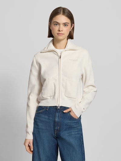 Frauenmodel trägt Slim Fit Strickjacke mit aufgesetzten Taschen von Tommy Jeans in Optik: Unifarben, Strickoptik, Label-Schriftzug, Reißverschluss, Stehkragen, langarm, gerippte Abschlüsse, Materialverarbeitung: Strick, Ganzkörperansicht von vorne.