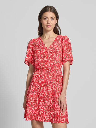 Frauenmodel trägt Leicht ausgestelltes Minikleid aus reiner Viskose Modell 'ALBA' von Vero Moda, rot mit weißem Punktmuster, V-Ausschnitt, Taillengürtel, kurzarm, Mini-Länge, ganzkörperansicht von vorne.