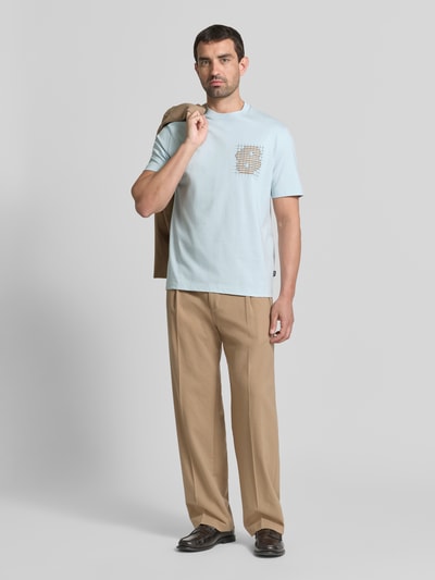 Mężczyzna nosi Męski t-shirt Regular Fit T-Shirt aus reiner Baumwolle model 'C-Tigerman 49' von BOSS w jasnoniebieskim kolorze z graficznym wzorem, brązowe spodnie, zdjęcie całej sylwetki z przodu.