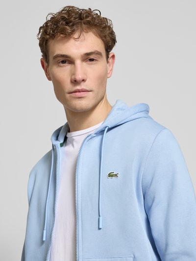 Męski model nosi Regular Fit Sweatjacke aus Baumwoll-Mix od Lacoste, jasnoniebieski, logo, zamek, długie rękawy, kaptur, bluza, bawełna-mix, krój regular fit, widok z przodu na górną część ciała.