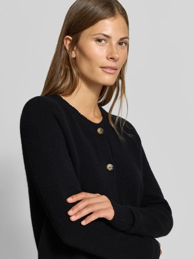 Frauenmodel trägt Strickjacke aus reinem Kaschmir von MYON in Schwarz mit Rundhalsausschnitt und Knopfleiste, Nahaufnahme Oberkörper. Die Strickjacke zeigt eine gerade Passform und feine Stricktextur. Rückenlänge und Ärmellänge bei Größe 36 sind 56 cm bzw. 66 cm.