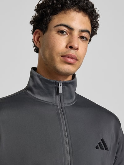 Männermodel trägt Slim Fit Trainingsanzug mit Logo-Stitching von adidas Sportswear in Grau mit Stehkragen, Reißverschluss, Nahaufnahme Oberkörper.