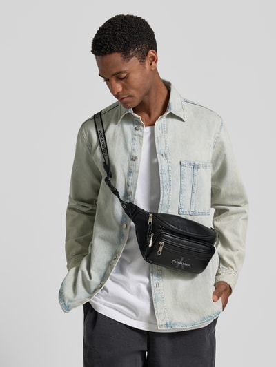 Männermodel trägt schwarze Umhängetasche mit Logo-Prägung Modell 'BOLD LOGO WEBBING WAISTBA' von CK Calvin Klein, Herren, Nahaufnahme Oberkörper.