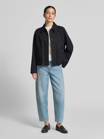 Modelka nosi Relaxed Fit Trenchcoat aus reiner Baumwolle od Tommy Hilfiger, efekt: jednolity kolor, krój relaxed fit, wykładany kołnierz, zapięcie: listwa guzikowa, długość: krój krótki, detal z logo, zakończenie rękawów: prosty, funkcja: hydrofobowy, wykończenie materiału: splot twill, materiał: bawełna, czarny, prostokątne kieszenie, tekstura gładka, Ganzkörperansicht von vorne.