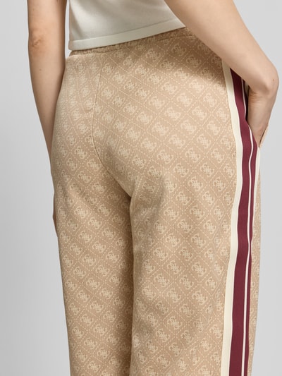 Frauenmodel trägt Wide Fit Sweatpants mit Allover-Logo-Print Modell 'Jacquard' von Guess Activewear, beige mit roten Streifen, Detailaufnahme Rückansicht. Material-Mix mit Baumwoll-Anteil, elastischer Bund, Eingrifftaschen, gemusterte Textur.