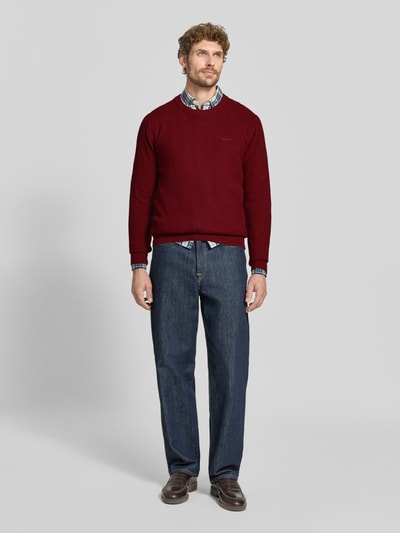 Männermodel trägt Regular Fit Pullover aus Baumwoll-Woll-Mix von GANT in Dunkelrot, kombiniert mit Jeans, Ganzkörperansicht von vorne.