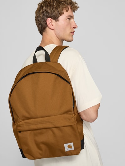 Männermodel trägt Rucksack mit Label-Patch Modell 'Jake' von Carhartt Work In Progress in bruin met zwarte details, buitenvak, draagband, verstelbare schouderbanden, hoofdvak met tweewegritssluiting, binnenvak van mesh met ritssluiting, breedte 40 cm, lengte 45 cm, diepte 14 cm, lengte schouderband 68 cm, driekwartaanzicht van voren