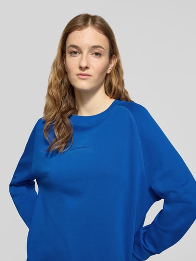 Modelka kobieca nosi Regular Fit Sweatshirt aus reiner Baumwolle od Marc O'Polo Denim, kolor jednolity, materiał bawełna, krój regular fit, nadruk z logo, okrągły dekolt, długi rękaw, mankiety i dół ze ściągaczem, tekstura typu sweat, z bocznymi rozcięciami, niebieski kolor; zbliżenie na górną część ciała z przodu.