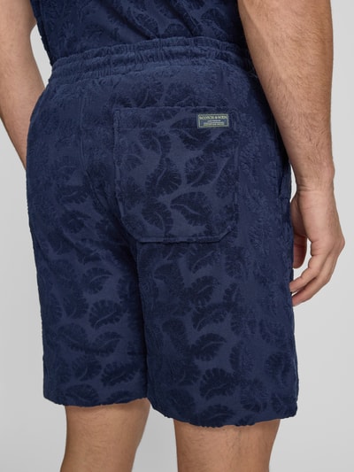 Männermodel trägt Relaxed Fit Shorts aus Frottee mit Gesäßtasche von Scotch & Soda, mieszanka bawełny, krój relaxed fit, frota, elastyczny pas, wpuszczane kieszenie, kieszeń z tyłu, detal z logo, szerokość w rozmiarze M: 41 cm, długość wewnętrzna nogawki w rozmiarze M: 18 cm, granatowy kolor, faktura froty, motyw liści, Ganzkörperansicht Rückansicht.