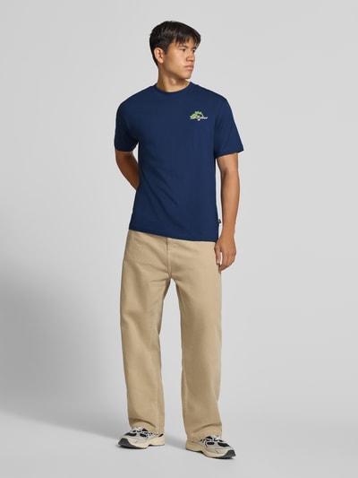 Männermodel trägt Regular Fit T-Shirt aus reiner Baumwolle mit Label-Detail von New Balance in Dunkelblau, Ganzkörperansicht von vorne. Modell 'GRAB A BITE TOKYO', Rückenlänge bei Größe M 70 cm.