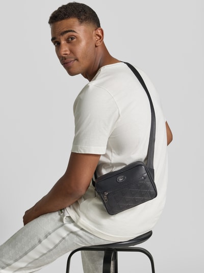 Männermodel trägt czarną torbę na ramię z nadrukiem monogramm 'Umhängetasche mit Monogramm-Print' od Lacoste, kontekst: męska torba na ramię, Ganzkörperansicht Rückansicht.