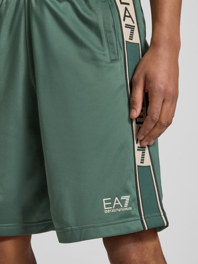 Mężczyznamodel nosi Szorty mit Logo und Eingrifftaschen von EA7 Emporio Armani, krój relaxed fit, elastyczny pas, logo, wpuszczane kieszenie, szerokość w rozmiarze M: 42 cm, długość wewnętrzna nogawki w rozmiarze M: 28 cm, detaliczny widok uda, zieleń, matowa tekstura.