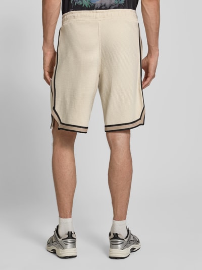 Männermodel trägt Shorts mit Strukturmuster und Label-Stitching von PEGADOR in Beige mit schwarzen Details, Reine Baumwolle, Regular-Fit, Eingrifftaschen, elastischer Bund, Bundweite 40 cm, Innenbeinlänge 26 cm, Ganzkörperansicht Rückansicht.