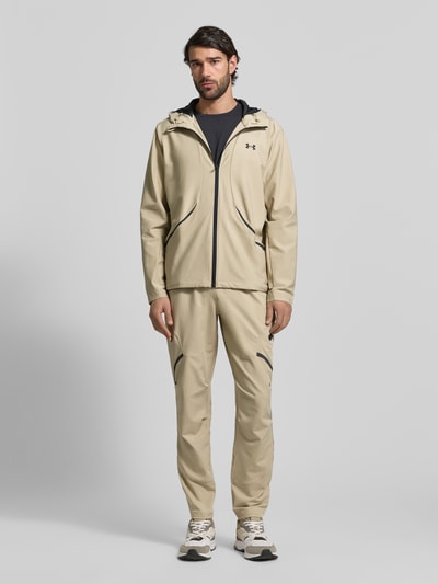 Männermodel trägt Regular Fit Jacke mit Logo-Print Modell 'Unstoppable' von Under Armour im Elastischer-Material-Mix, Reißverschluss, Eingrifftaschen, Kapuze mit Stehkragen, in Beige mit glatter Textur, Ganzkörperansicht von vorne.