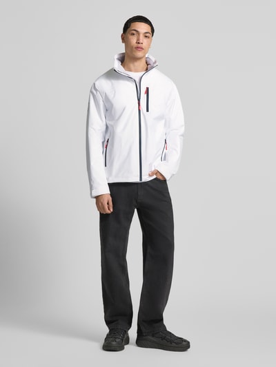 Männermodel trägt weiße Midlayer Jacke mit Stehkragen Modell 'CREW' von Helly Hansen, Gerader-Schnitt, Logo-Print, Reißverschlusstaschen, Ganzkörperansicht von vorne.