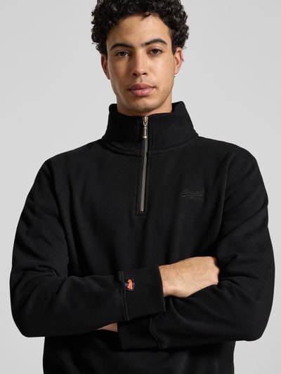 Männermodel trägt Regular Fit Sweatshirt mit Logo-Stitching von Superdry aus schwarzem Baumwoll-Mix, Nahaufnahme Oberkörper. Rückenlänge 65 cm, Ärmellänge 63 cm, Stehkragen mit Reißverschluss, elastische Abschlüsse.