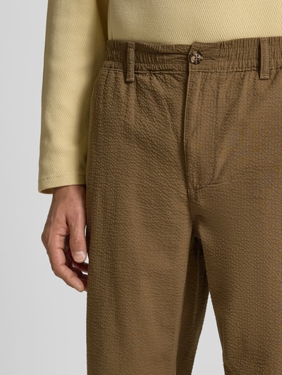 Männermodel trägt Stoffhose aus reiner Baumwolle von Forét, Strukturmuster, elastischer Bund mit Knopfverschluss, Eingriff- und Gesäßtaschen, Relaxed-Fit, braune Farbe, Nahaufnahme Oberkörper.