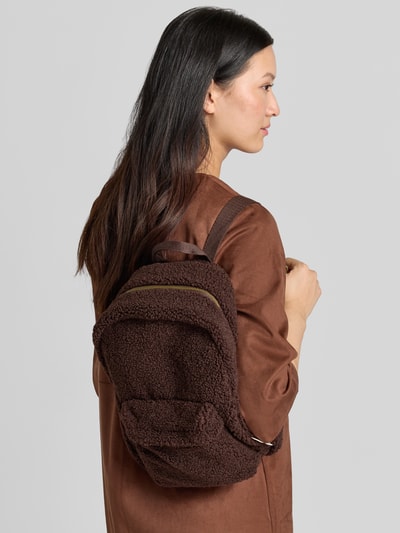 Frauenmodel trägt Rucksack aus Teddyfell von Studio Noos in Dunkelbraun mit goldfarbenem Reißverschluss, Ganzkörperansicht Rückansicht. Hauptfach, Außen- und Innenfach mit Reißverschluss, verstellbare Schulterriemen, Maße: 35x39x15 cm, Schulterriemen 50 cm.