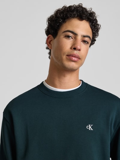 Männermodel trägt Sweatshirt aus reiner Baumwolle von Calvin Klein Jeans in Dunkelgrün mit Label-Stitching, Rundhalsausschnitt, Nahaufnahme Oberkörper.