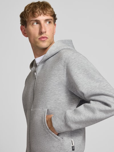Männermodel trägt Relaxed Fit Sweatjacke mit Zweiwege-Reißverschluss von JACK & JONES in Grau, strukturierte Oberfläche, Nahaufnahme Oberkörper. Elastischer Material-Mix, getrennte Känguru-Tasche, elastische Abschlüsse.