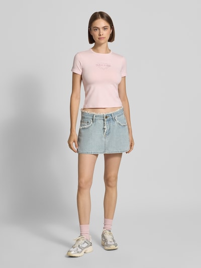 Frauenmodel trägt Slim Fit T-Shirt mit Strasssteinbesatz Modell 'Heart Baby' von GUESS Jeans in Hellrosa und kurzem Jeansrock, Ganzkörperansicht von vorne.