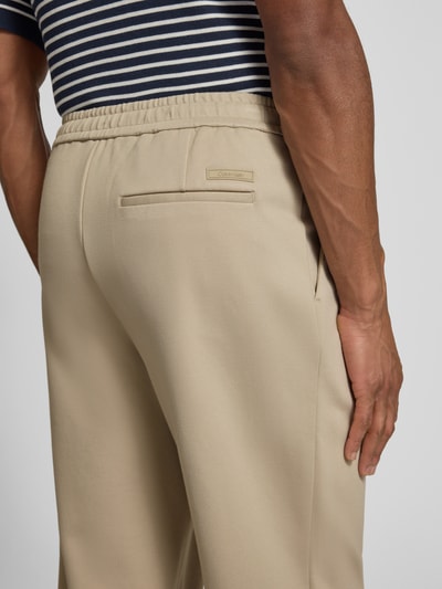 Männermodel trägt Hose mit französischen Eingrifftaschen von CK Calvin Klein, beigefarbener Baumwoll-Mix, elastischer Bund, Nahaufnahme Rückansicht.