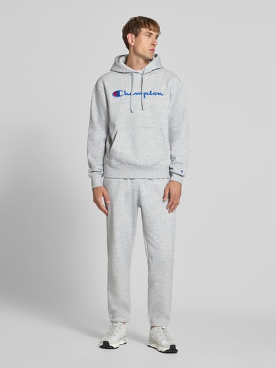 Männermodel trägt Hoodie mit Label-Stitching von Champion in Hellgrau, lockerer Schnitt, Kängurutasche, Kapuze, Baumwoll-Mix, Ganzkörperansicht von vorne.