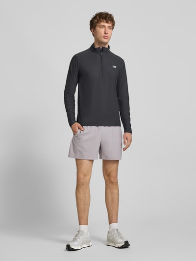 Męski model nosi Regular Fit Sweatshirt mit Troyer-Kragen od New Balance, czarny kolor, elastyczna mieszanka materiałów, nadruk z logo, długość tyłu w rozmiarze M: 69 cm, widok całej sylwetki z przodu.