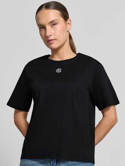 Kobieta nosi Regular Fit T-Shirt aus reiner Baumwolle Model 'Melea' od MSCH Copenhagen, czarny, nadruk z logo, okrągły dekolt, damski t-shirt, damski, kontekst: damski t-shirt z bawełny, krój regular fit, nadruk z logo, okrągły dekolt, długość tyłu w rozmiarze XS/S: 57 cm, Ganzkörperansicht z przodu.
