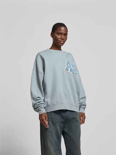 Frauenmodel trägt Oversized Sweatshirt aus reiner Baumwolle von Anine Bing in Hellblau mit Label-Detail, elastische Abschlüsse, Rundhalsausschnitt, Rückenlänge bei Größe S 64 cm, Ganzkörperansicht von vorne.