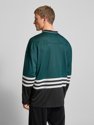 Männermodel trägt Longsleeve mit Label-Detail von adidas Originals, grün-schwarz, Lochstickerei, Lockerer-Schnitt, Ganzkörperansicht Rückansicht.