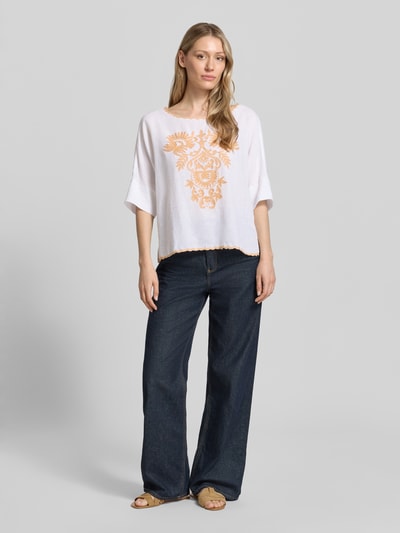 Frauenmodel trägt Blusenshirt mit Leinen-Anteil und Motiv-Stitching von Christian Berg Woman, weiß mit orangem Motiv, Loose-Fit, U-Boot-Ausschnitt, Viskose-Mix, Rückenlänge 55 cm, Ärmellänge 34 cm, Ganzkörperansicht von vorne.